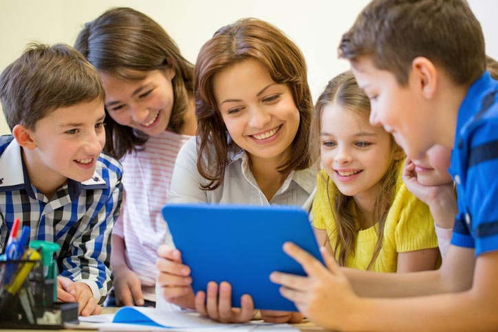 Eine kleine Gruppe Kinder lernt Englisch und schaut sich etwas auf einem Tablett an. Die Gruppe steht um die Englisch-Lehrerin herum.